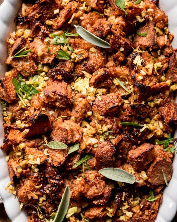 An up close overhead shot shows a dish of baked vegan stuffing with rustically torn sourdough bread, balsamic shallots, celery, and herbs. Whole sage leaves garnish the top.