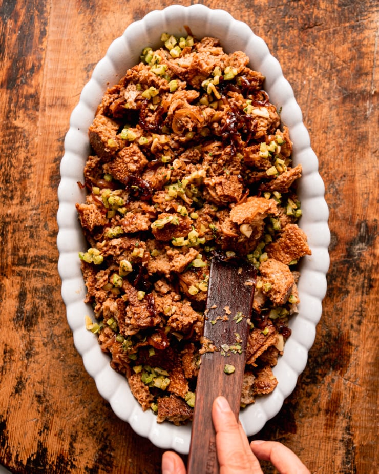 An overhead shot shows a hand using a wooden utensil to distribute stuffing in a baking dish.
