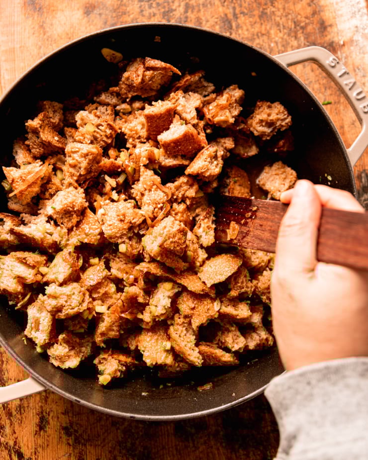 An overhead shot shows a hand using a wooden utensil to stir up bread pieces, shallots, herbs, and celery together in a braiser-style pot.