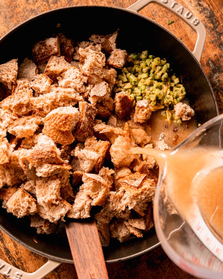 An overhead shot shows a vegetable stock mixture being poured into a pot with torn bread pieces, chopped celery, shallots, and herbs.