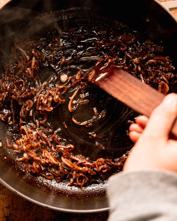 An overhead shot shows a hand using a wooden utensil to stir up balsamic shallots in a braiser-style pot.