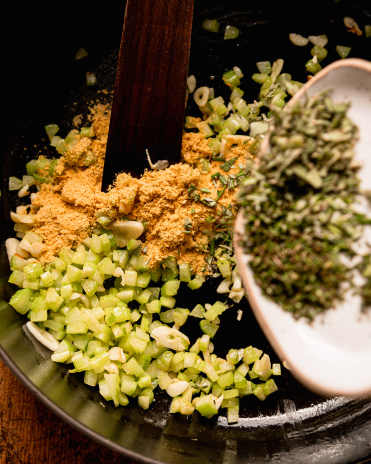 An overhead shot shows chopped herbs being added to a pot with sautéed celery, garlic, and some nutritional yeast.