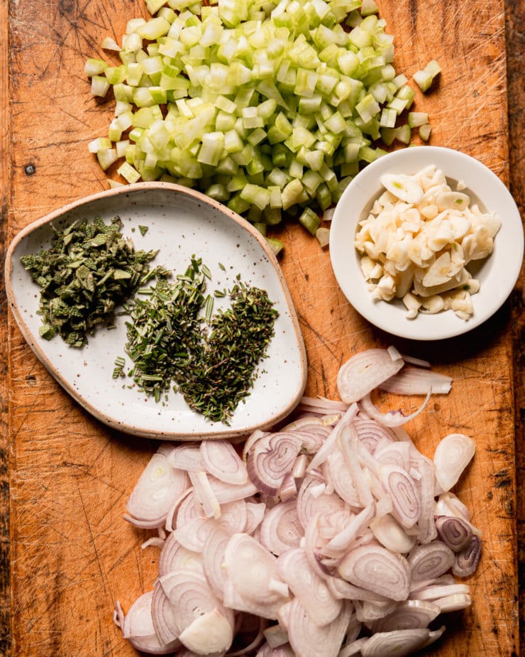 An overhead shot shows diced celery, sliced garlic, sliced shallots, and chopped herbs; all on a worn wooden cutting board.