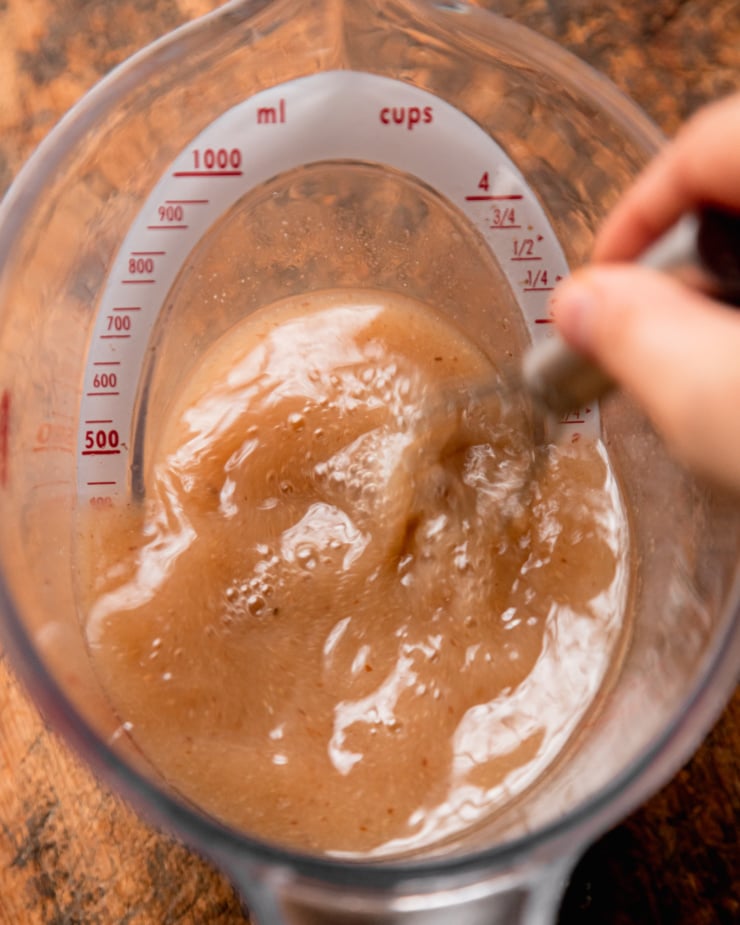 An overhead shot shows a hand using a whisk to combine vegetable stock with miso and ground flaxseed in a large liquid measuring cup.
