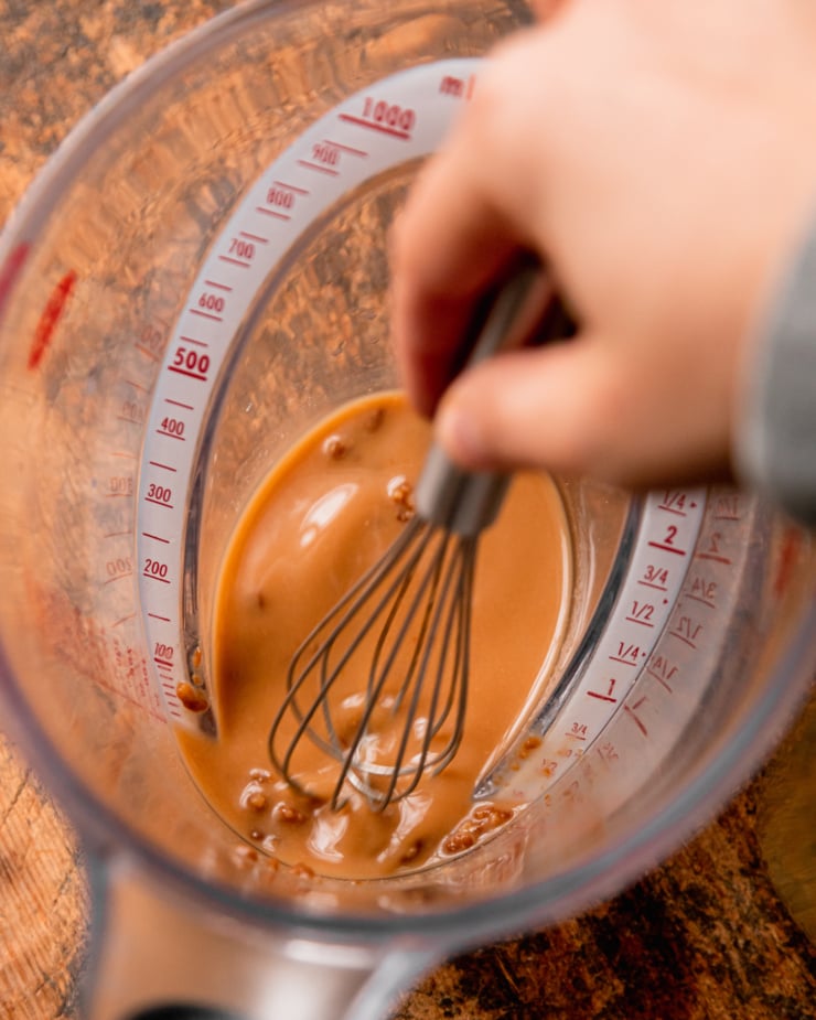 An overhead shot shows a hand using a whisk to mix miso paste into a bit of vegetable stock in a large measuring cup.