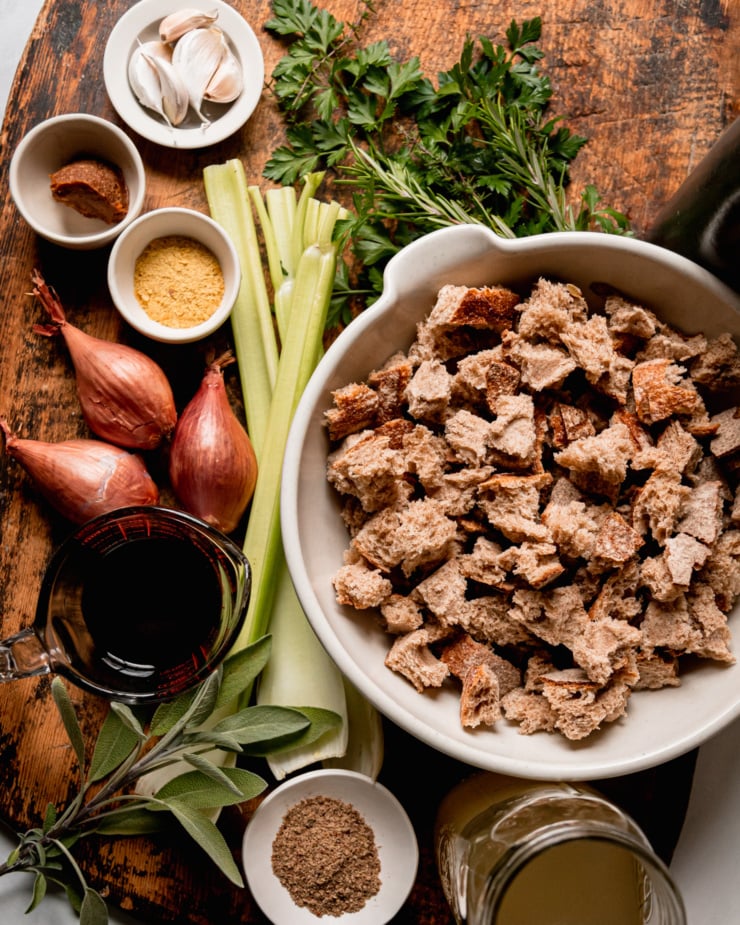 An overhead shot shows ingredients used in a vegan stuffing recipe: parsley, rosemary, thyme, sage, torn sourdough bread, vegetable stock, ground flaxseed, celery, balsamic vinegar, shallots, nutritional yeast, miso, and garlic.