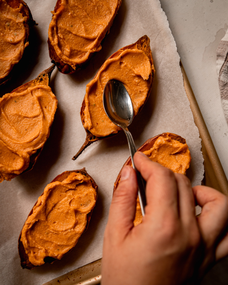 An overhead shot shows a hand swooping a sweet potato filling that has gone into baked sweet potatop halves.