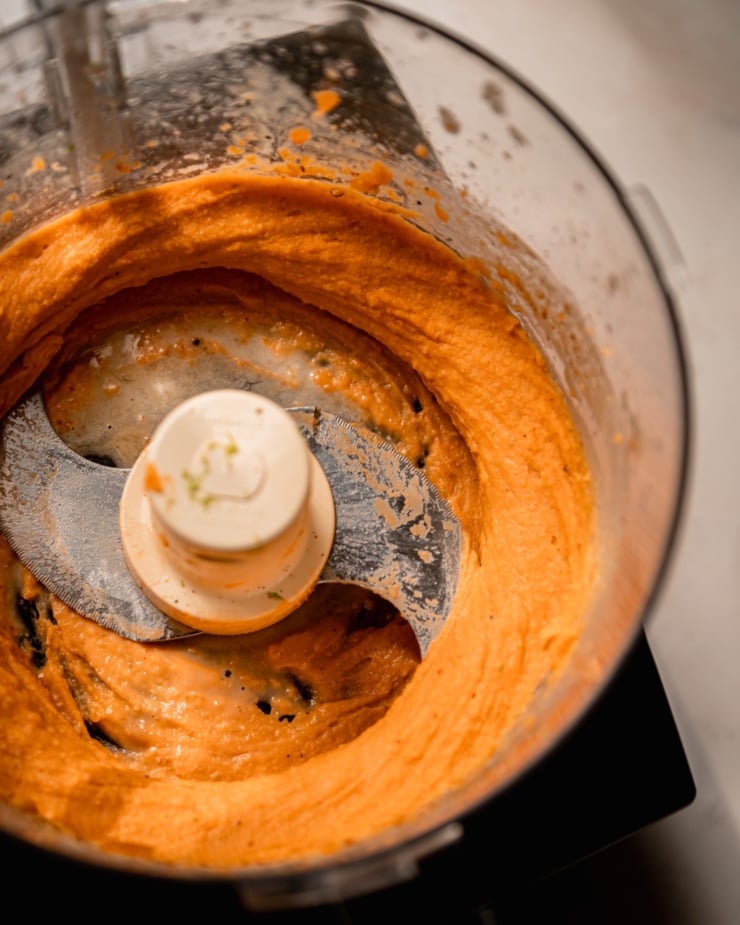An overhead shot shows a whipped sweet potato mixture in a food processor that also contains tahini, lime, olive oil, spices, and maple syrup.
