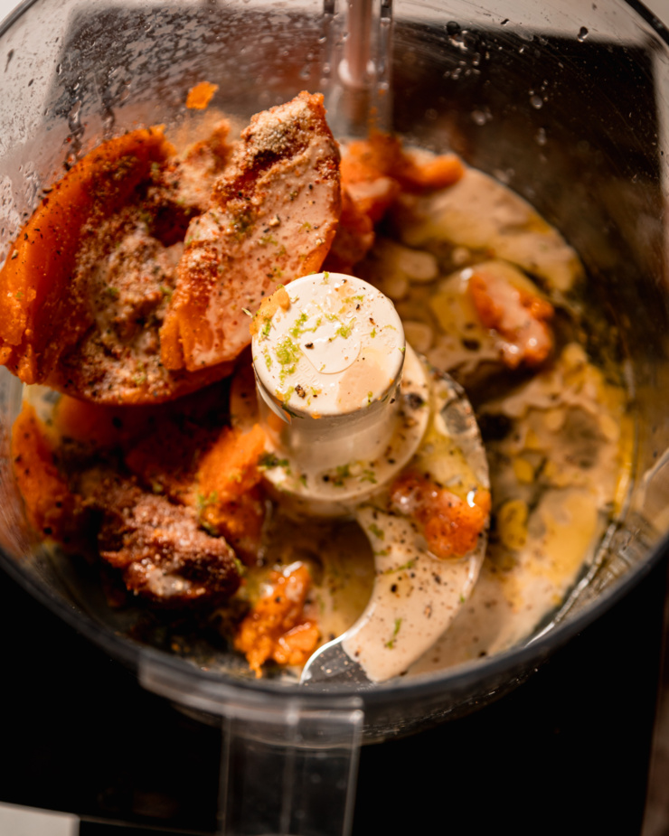 An overhead shot shows some ingredients in a food processor: cooked sweet potato, lime zest, lime juice, maple syrup, miso, maple syrup, and spices.