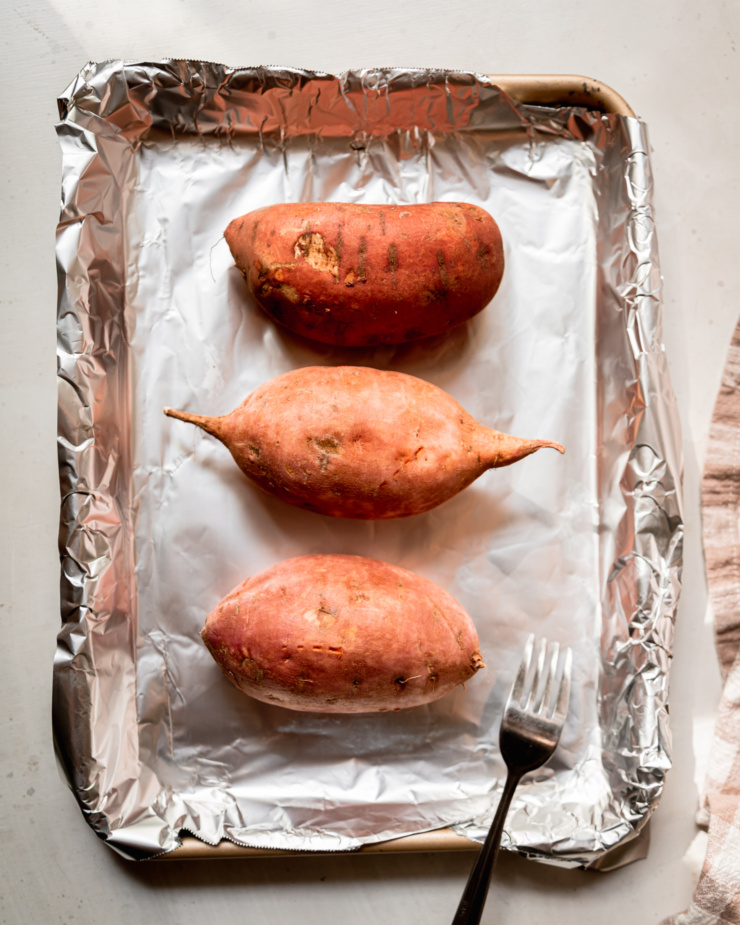 An overhead shot shows whole sweet potatoes on a foil-lined baking sheet with holes pricked into them.