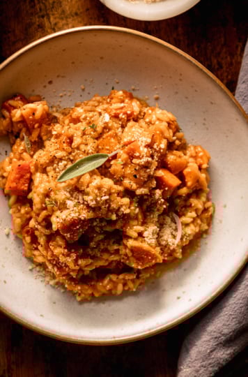 An overhead shot shows a single serving of vegan butternut squash risotto in a wide serving bowl. The risotto is dusted with vegan "parmesan" and topped with a couple fresh sage leaves.