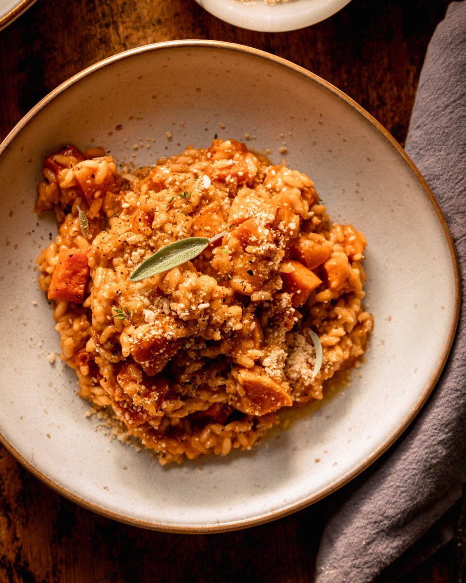 An overhead shot shows a single serving of vegan butternut squash risotto in a wide serving bowl. The risotto is dusted with vegan "parmesan" and topped with a couple fresh sage leaves.