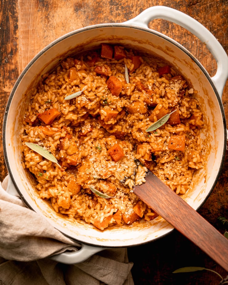 An overhead shot shows a creamy risotto in a pot with chunks of butternut squash. The risotto is in a Dutch oven-style pot with a wooden utensil sticking out.