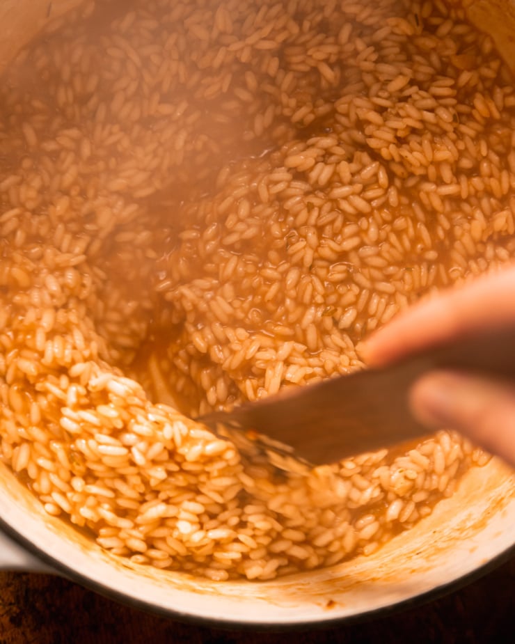 An up close, overhead shot shows a hand using a wooden utensil to stir arborio rice, vegetable stock, and seasonings. There is steam rising up from the rice.