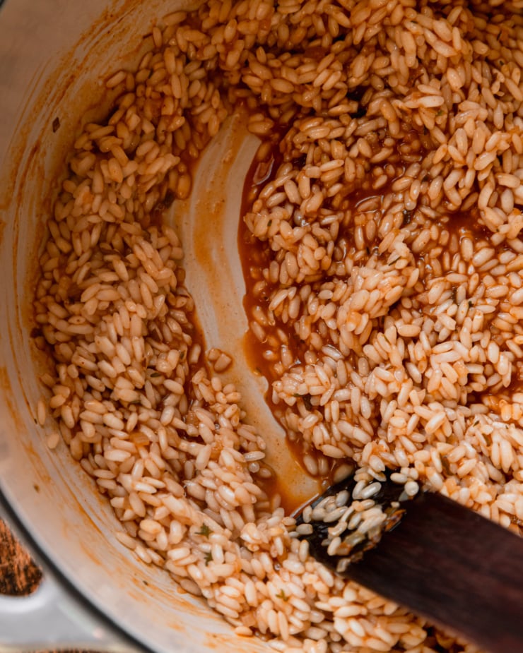 An overhead shot shows a "track" made by a wooden utensil in a pot of arborio rice, vegetable stock, and seasonings.