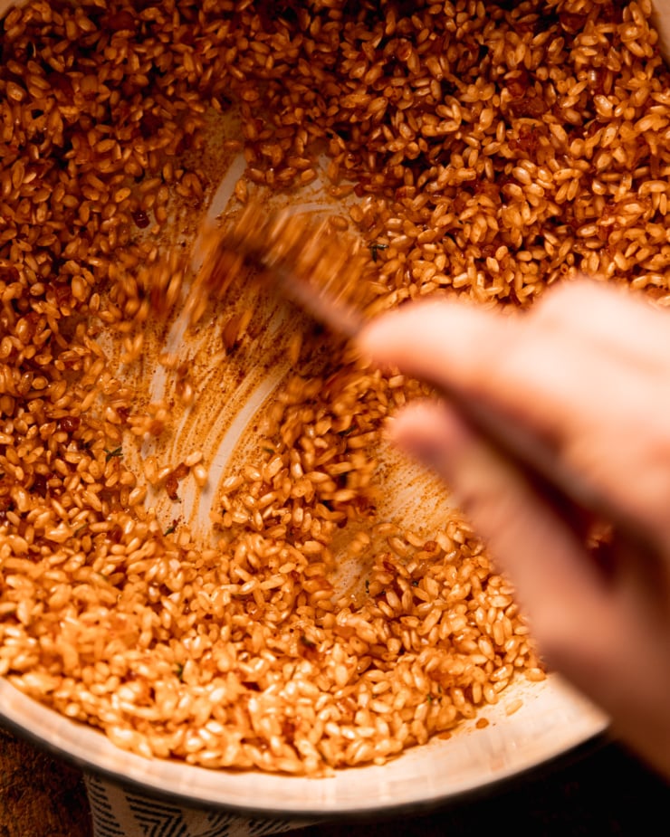 An overhead shot shows a hand using a wooden utensil to toast arborio rice in olive oil and seasonings.