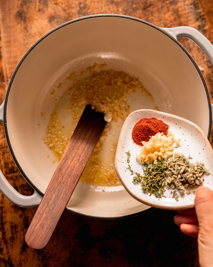 An overhead shot shows sautéed shallots in a Dutch oven. A hand is emptying a plate with minced sage and thyme, paprika, and minced garlic into the pot.