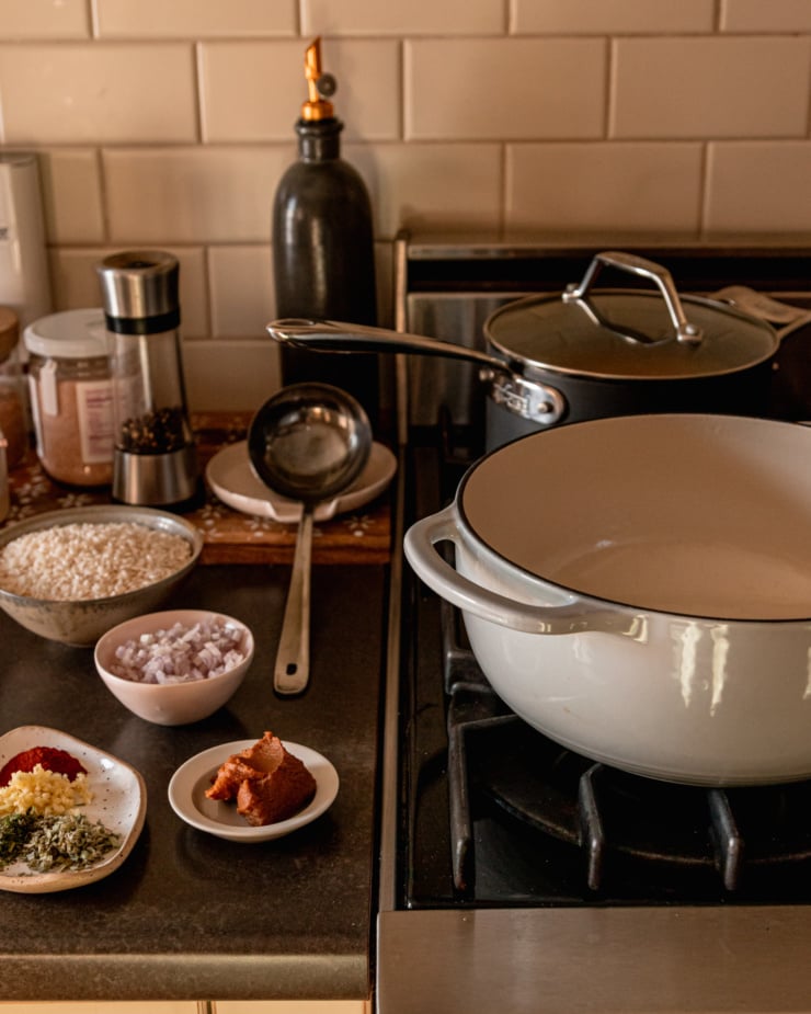A slight 3/4 angle image shows a stovetop with two pots and some cooking ingredients nearby: miso, diced shallots, minced garlic, spices, herbs, and arborio rice.
