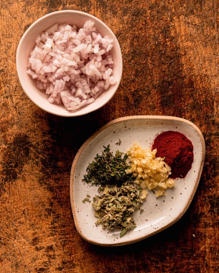 An overhead shot shows a small bowl with minced shallots and a small plate with spices, chopped herbs, and minced garlic.