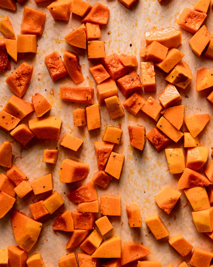 An overhead shot shows cubes of butternut squash coated in olive oil, salt, and pepper on a parchment-lined baking sheet.