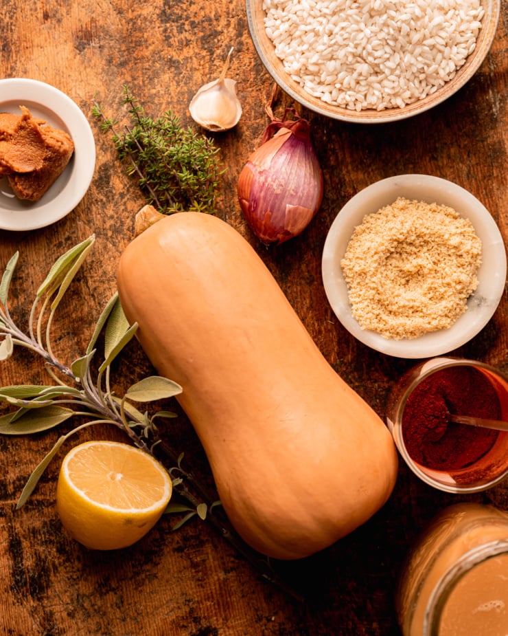 An overhead shot shows ingredients on a worn wood cutting board: a whole butternut squash, arborio rice in a bowl, a shallot, garlic clove, lemon, sage, thyme, miso, smoked paprika, vegetable stock, and a bowl of vegan "parmesan" sprinkle.