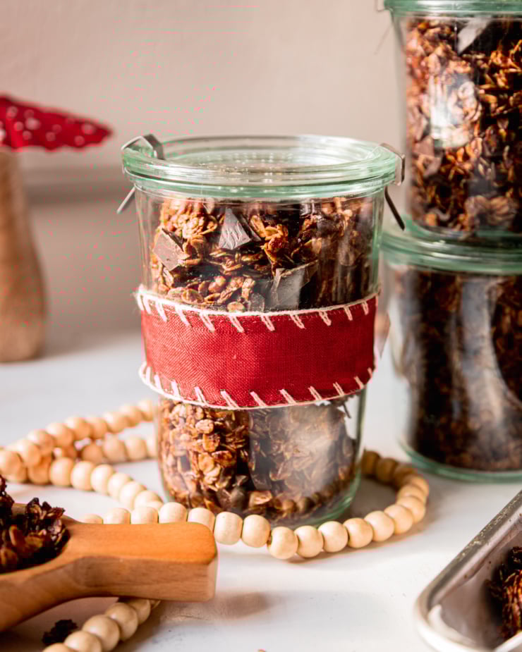 A head on shot shows a few jars of ginger molasses granola with dark chocolate chunks. One jar is wrapped in a red fabric ribbon.