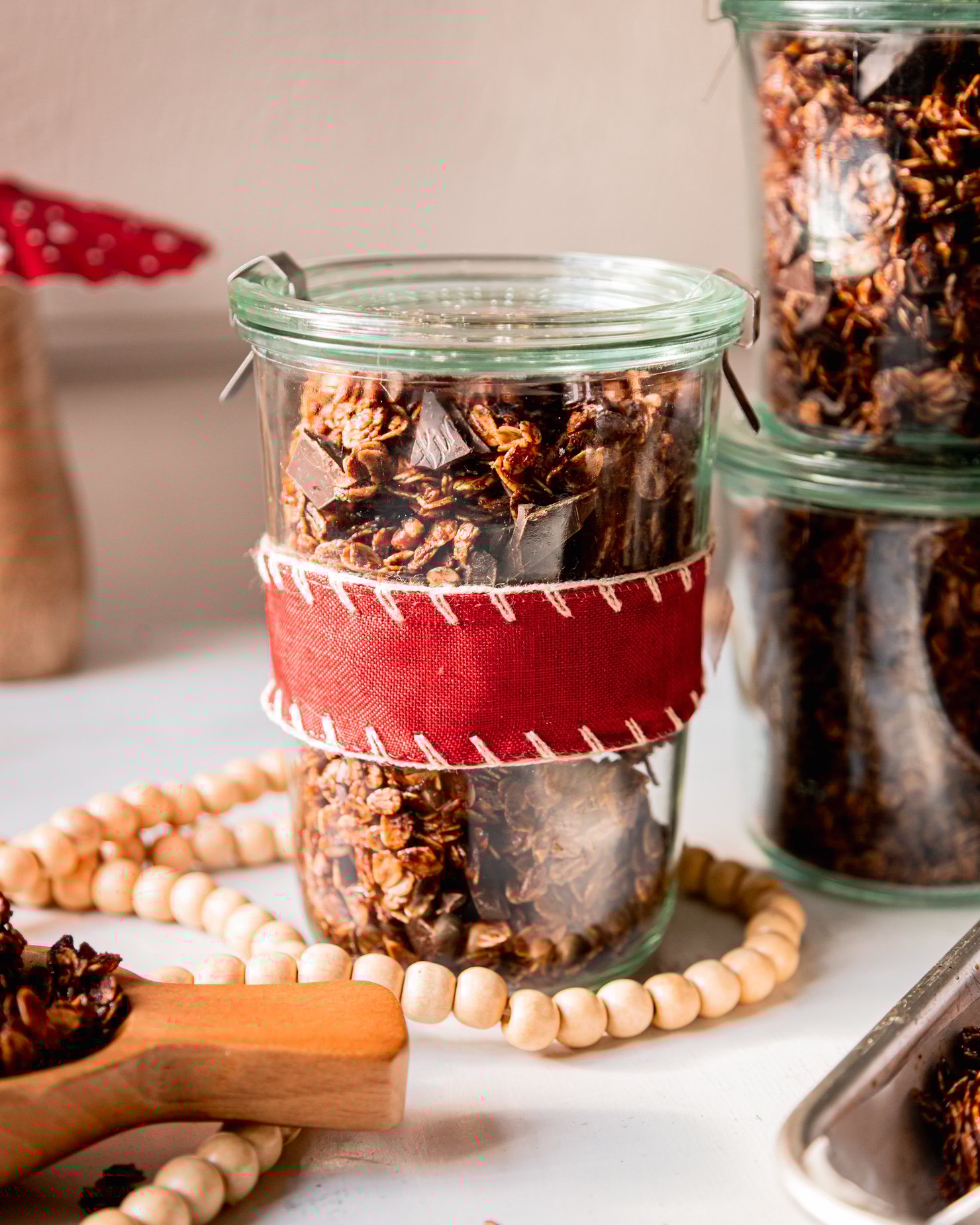 A head on shot shows a few jars of ginger molasses granola with dark chocolate chunks. One jar is wrapped in a red fabric ribbon.