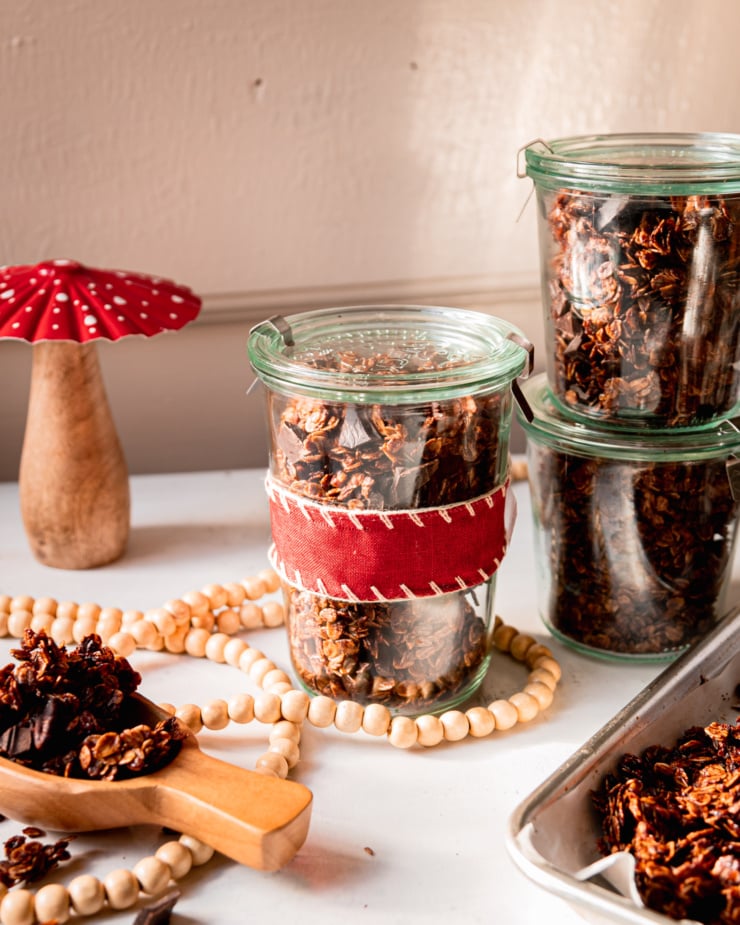 A head on shot shows a few jars of ginger molasses granola with dark chocolate chunks. One jar is wrapped in a red fabric ribbon. A decorative wood/metal mushroom is nearby.
