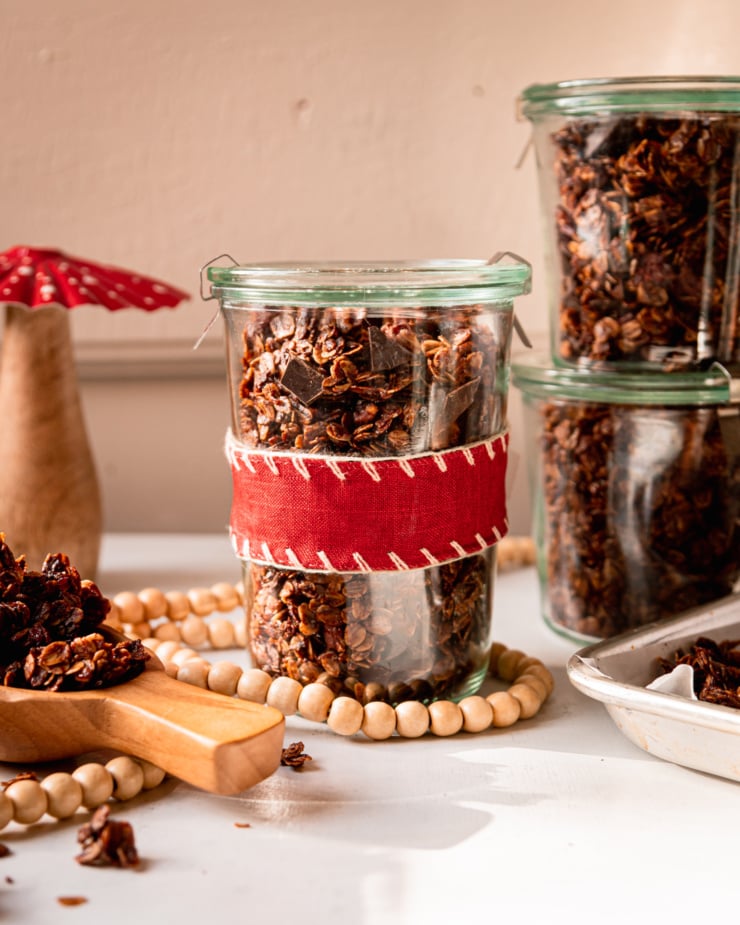 A head on shot shows a few jars of ginger molasses granola with dark chocolate chunks. One jar is wrapped in a red fabric ribbon.