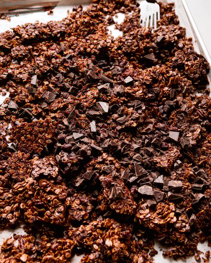 A 3/4 angle shot shows a baking sheet filled with ginger molasses granola. The granola is deep golden brown and features dark chocolate and dried cranberries.