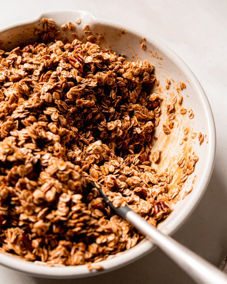 A 3/4 angle shot shows oats in a large bowl, coated in a maple syrup mixture with chopped pecans.