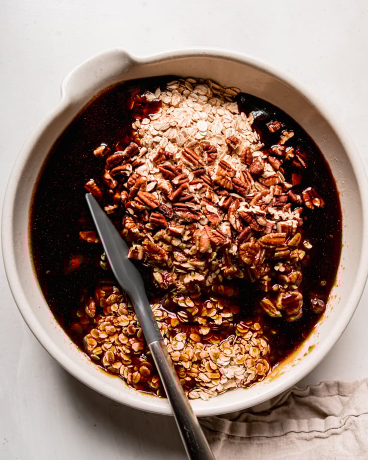 AN overhead shot shows a large bowl filled with toasted oats, chopped pecans, and a spiced maple syrup mixture. A spatula is sticking out of the bowl.