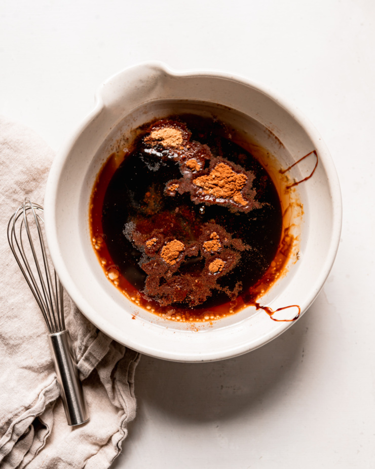 An overhead shot shows a medium bowl with a pour spout that is filled with molasses, maple syrup, spices, salt, and coconut oil. A whisk and napkin are nearby.