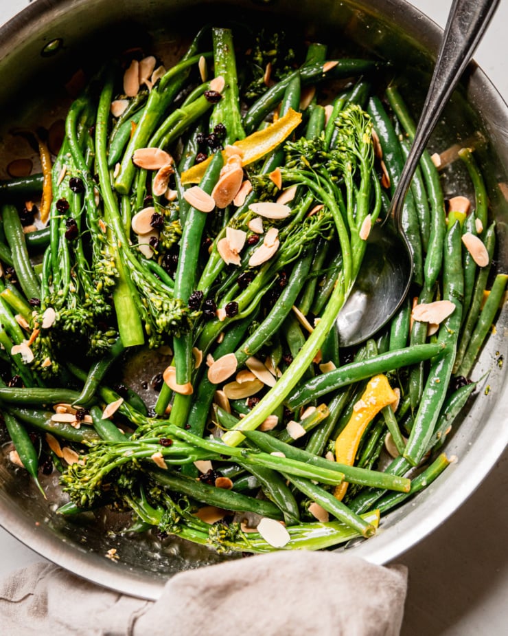 An overhead shot shows sautéed broccolini and green beans in a skillet. The vegetables are topped with toasted almond slices and soaked currants. A spoon is sticking out of the skillet.