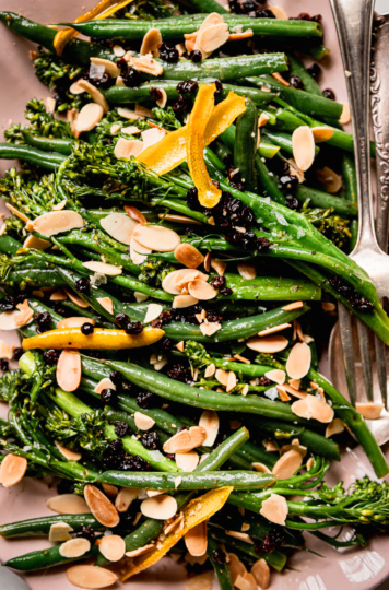 An up close, overhead shot shows sautéed broccolini and green beans on a platter. The vegetables are topped with soaked currants, toasted almond slices and strips of lemon peel.