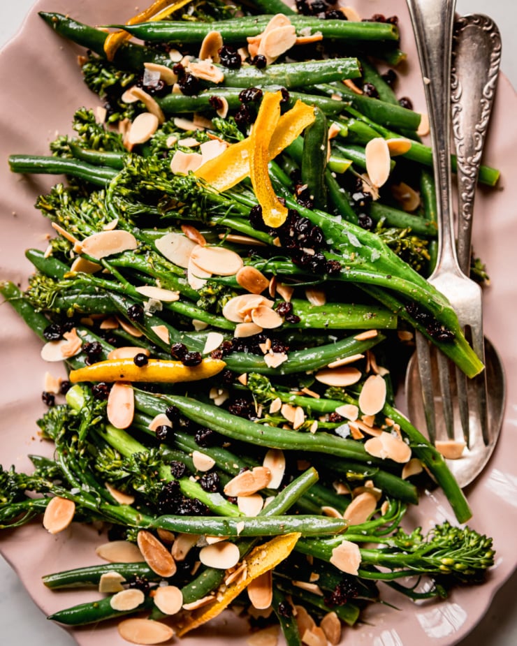 An up close, overhead shot shows sautéed broccolini and green beans on a platter. The vegetables are topped with soaked currants, toasted almond slices and strips of lemon peel.