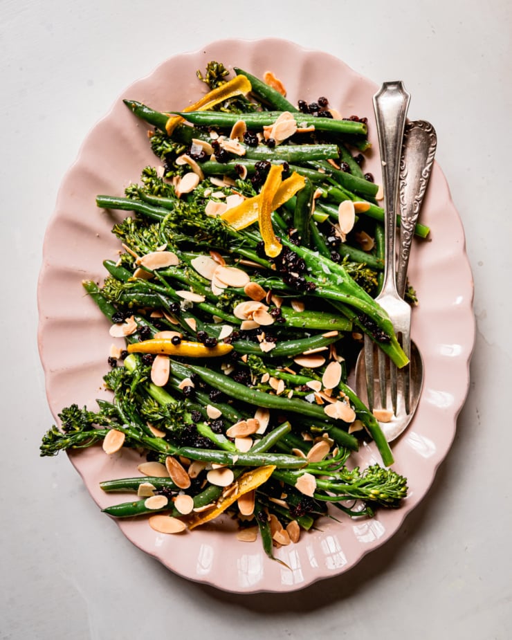 An overhead shot shows sautéed broccolini and green beans on a platter. The vegetables are topped with soaked currants, toasted almond slices and strips of lemon peel.