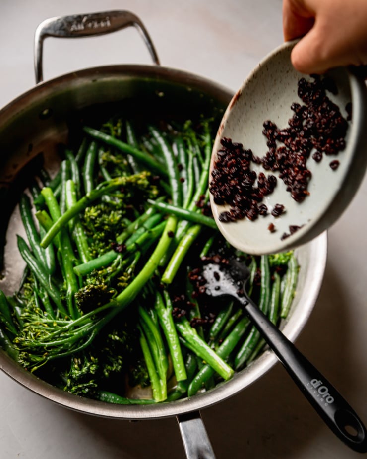 An overhead shot shows soaked currants being added to a skillet with green vegetables.