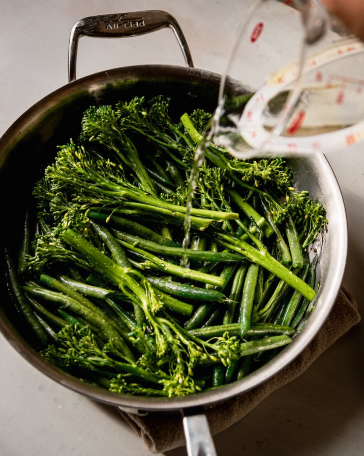 An overhead shot shows water being poured onto sautéed broccolini and green beans in a skillet.
