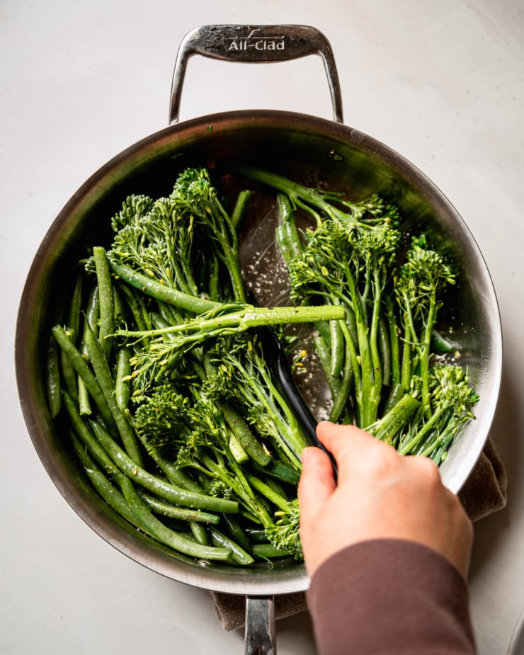 An overhead shot shows a hand using a spatula to stir up some green beans and broccolini in a skillet.