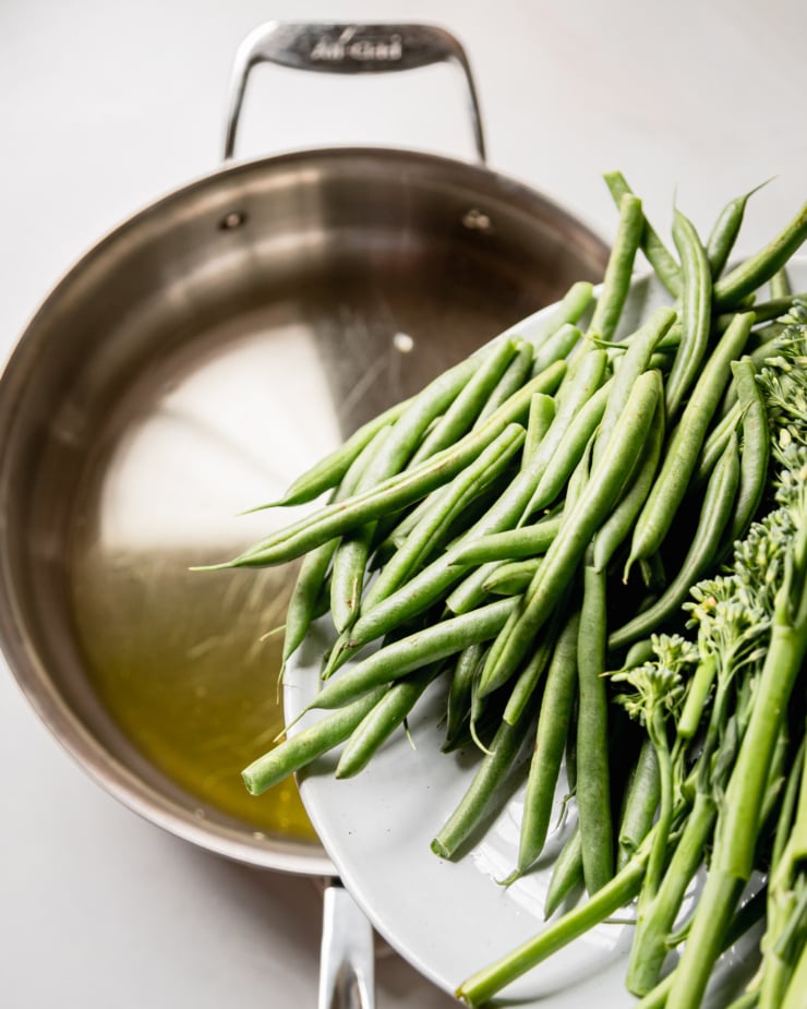 An overhead shot shows green beans and broccolini that are about to be added to a skillet with oil in it.