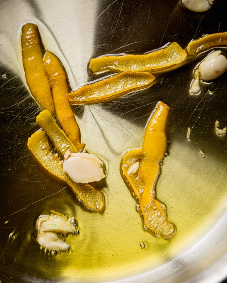 An up close, overhead shot shows lemon peels and smashed garlic cloves sizzling in hot oil.