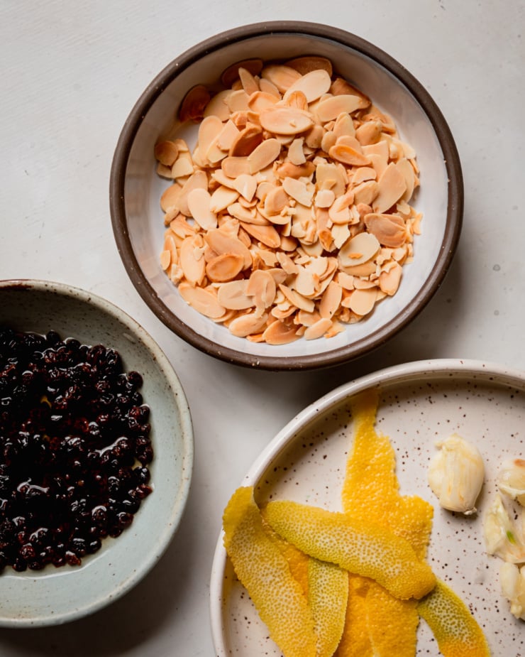 An overhead shot shows bowls of toasted lamonds and soaked currants. Strips of lemon peel and smashed garlic cloves are also seen on a plate.