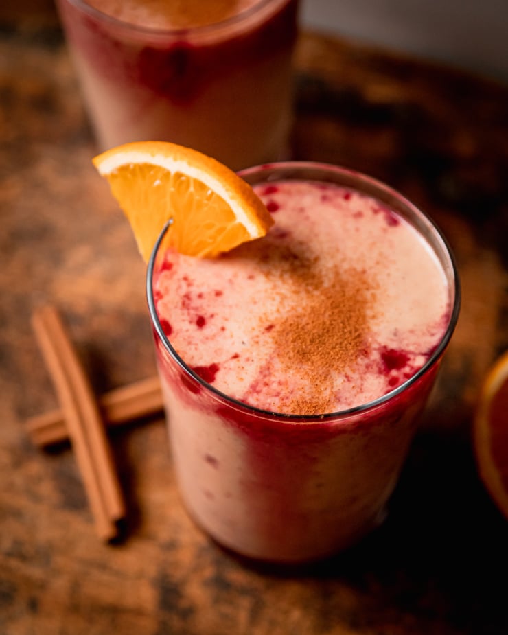 An overhead shot shows a glass filled with an orange cranberry smoothie with a little orange slice garnish. Cranberry juice "tie dye" stripes are visible on the surface.