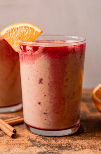 A head-on shot shows two glasses of orange cranberry smoothie, garnished with little orange slices on the rim.