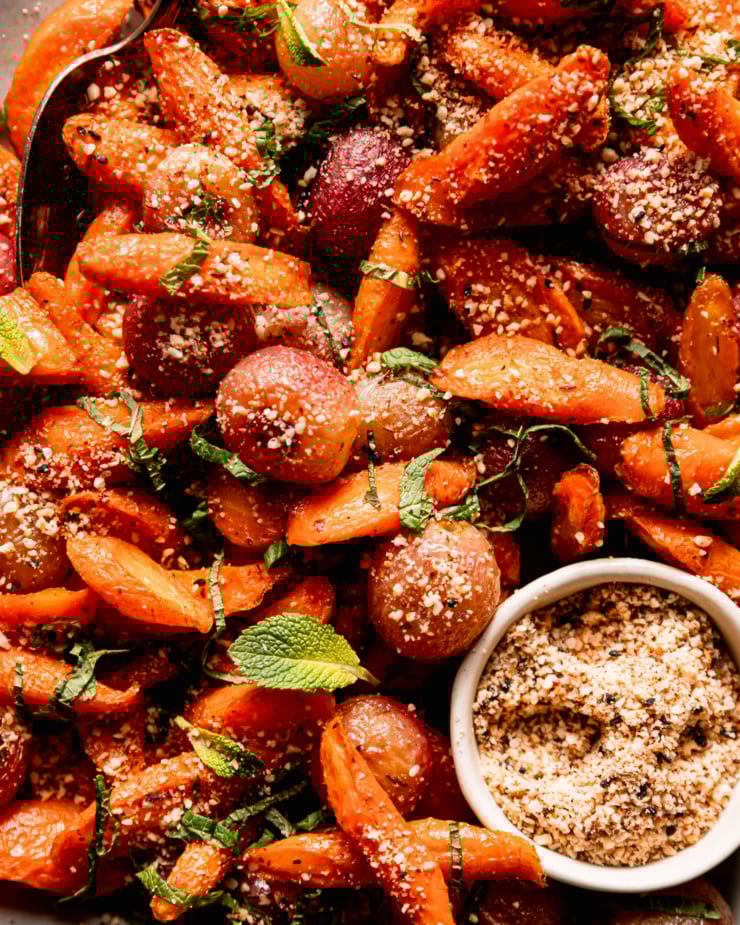 An up close, overhead shot shows a platter of oven roasted carrot side dish featuring roasted grapes, sprinkles of hazelnut-based dukkah, and finely sliced mint.