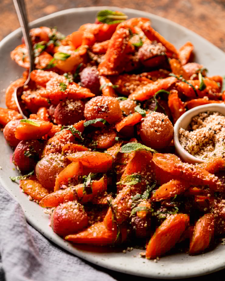 A 3/4 angle shot shows a platter of oven roasted carrot side dish featuring roasted grapes, sprinkles of hazelnut-based dukkah, and finely sliced mint.