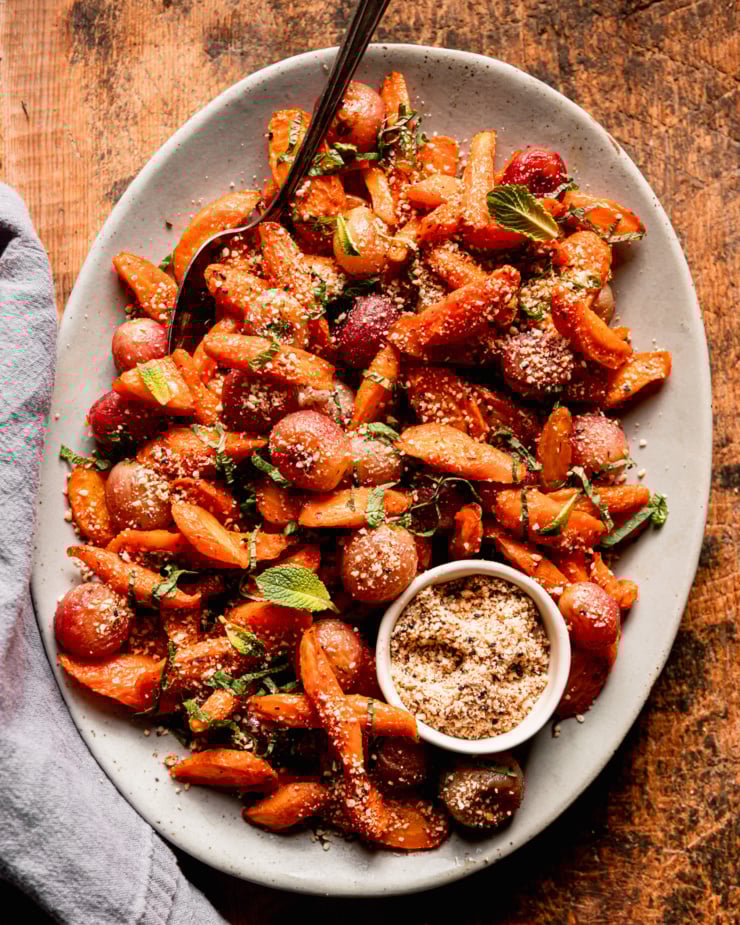 An overhead shot shows a platter of oven roasted carrot side dish featuring roasted grapes, sprinkles of hazelnut-based dukkah, and finely sliced mint.