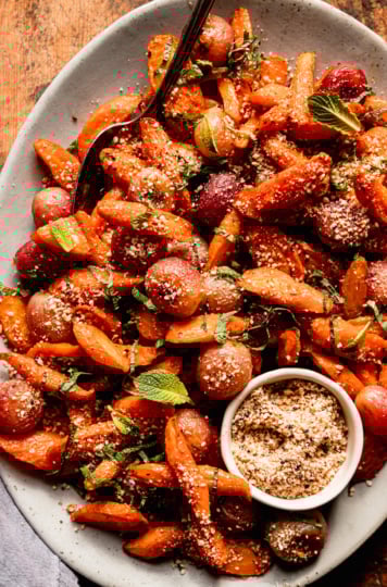An overhead shot shows a platter of oven roasted carrot side dish featuring roasted grapes, sprinkles of hazelnut-based dukkah, and finely sliced mint.
