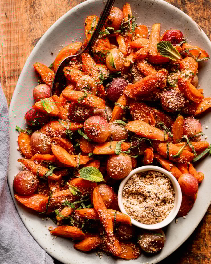 An overhead shot shows a platter of oven roasted carrot side dish featuring roasted grapes, sprinkles of hazelnut-based dukkah, and finely sliced mint.